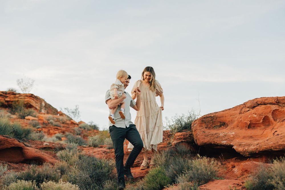 A couple stands in a scenic desert landscape, holding a young child, with red rock formations in the background.