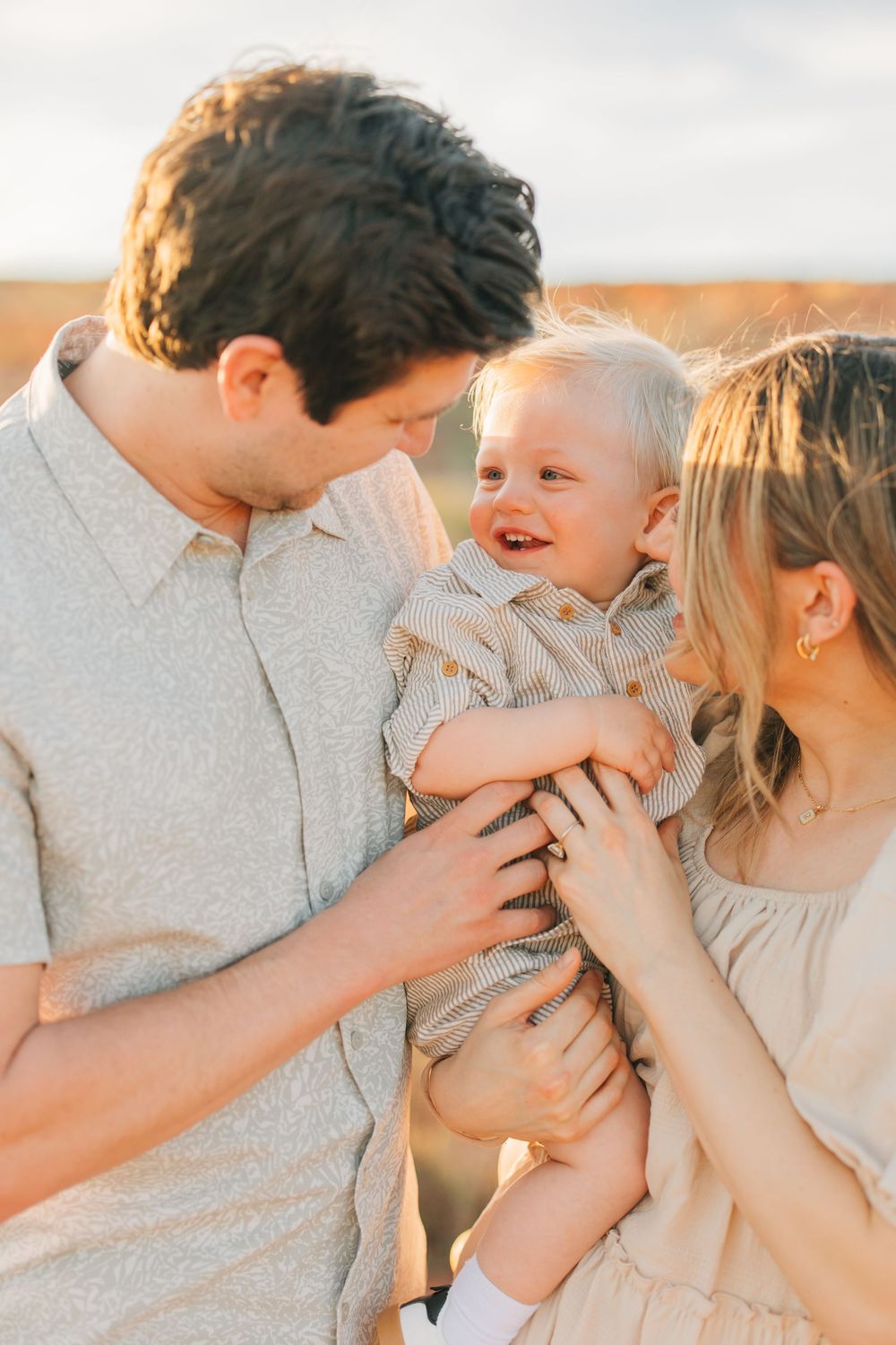 A man holding a smiling baby while a woman stands next to them, all in a scenic outdoor environment.