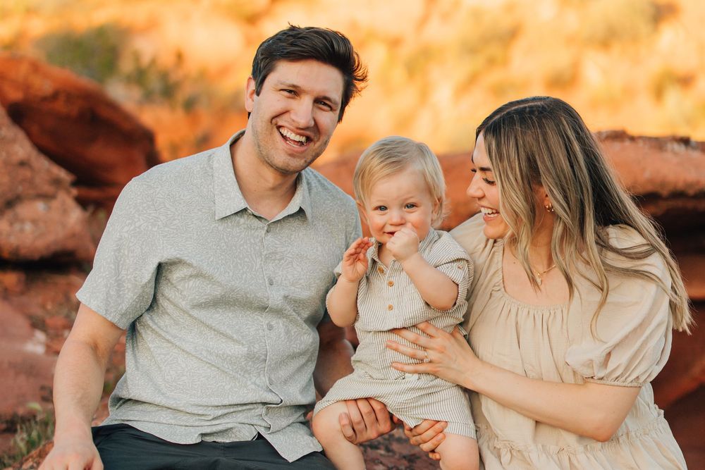 A happy family of three, with a father, mother, and young child, sitting outdoors in a natural setting.