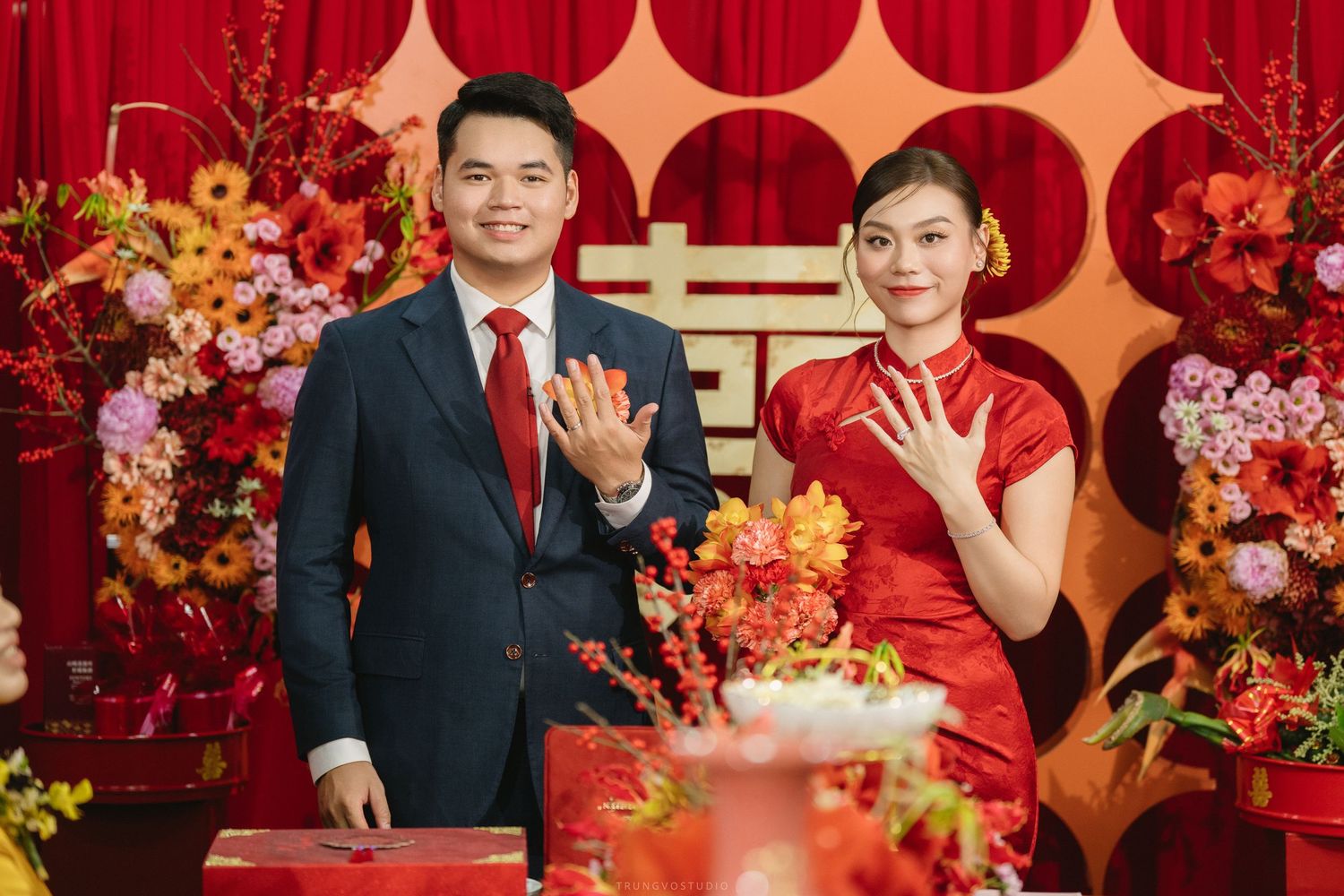 Couple in traditional Chinese wedding attire posing with red envelopes at engagement ceremony.