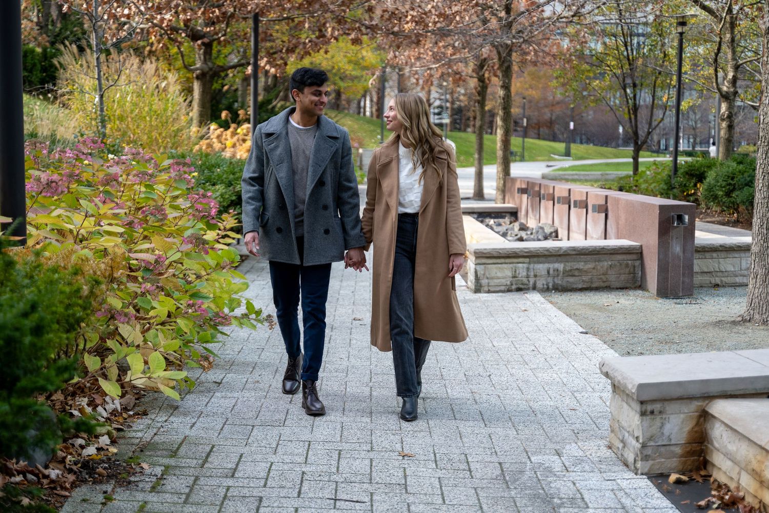 Two people walking together on a stone path through a garden or park area during autumn season.