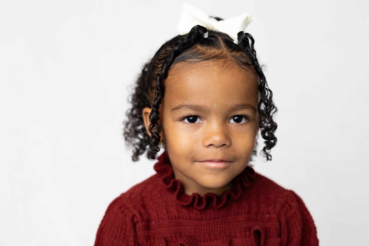 Child wearing a red sweater and hair bows poses for a close-up portrait.