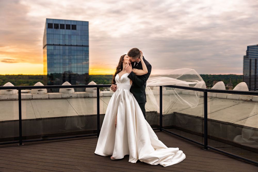 Bride and groom share a romantic embrace on a rooftop at sunset, with a flowing veil and city skyline backdrop.