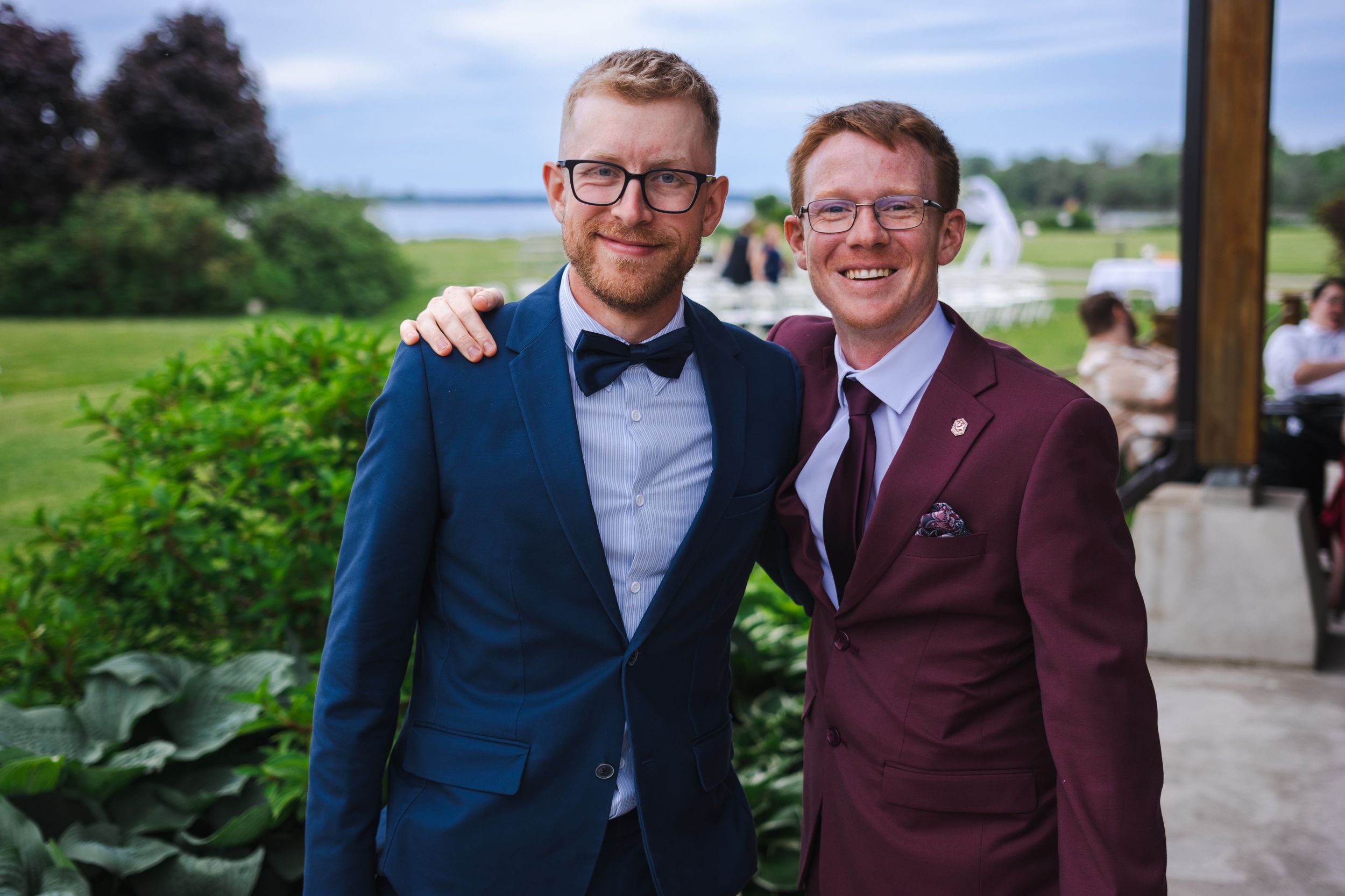 Two people in formal attire stand together outdoors, one wearing a navy suit and bow tie, the other in a burgundy suit.