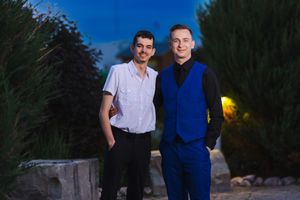 Two men stand together at dusk, one wearing a white shirt and the other in a blue suit.