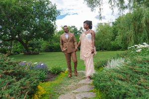 Two people walk along a garden path surrounded by lush greenery, colorful flowers and willow trees on a summer day.