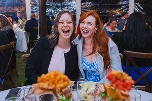 Two friends share a joyful laugh together at an outdoor reception with orange flowers on the table.