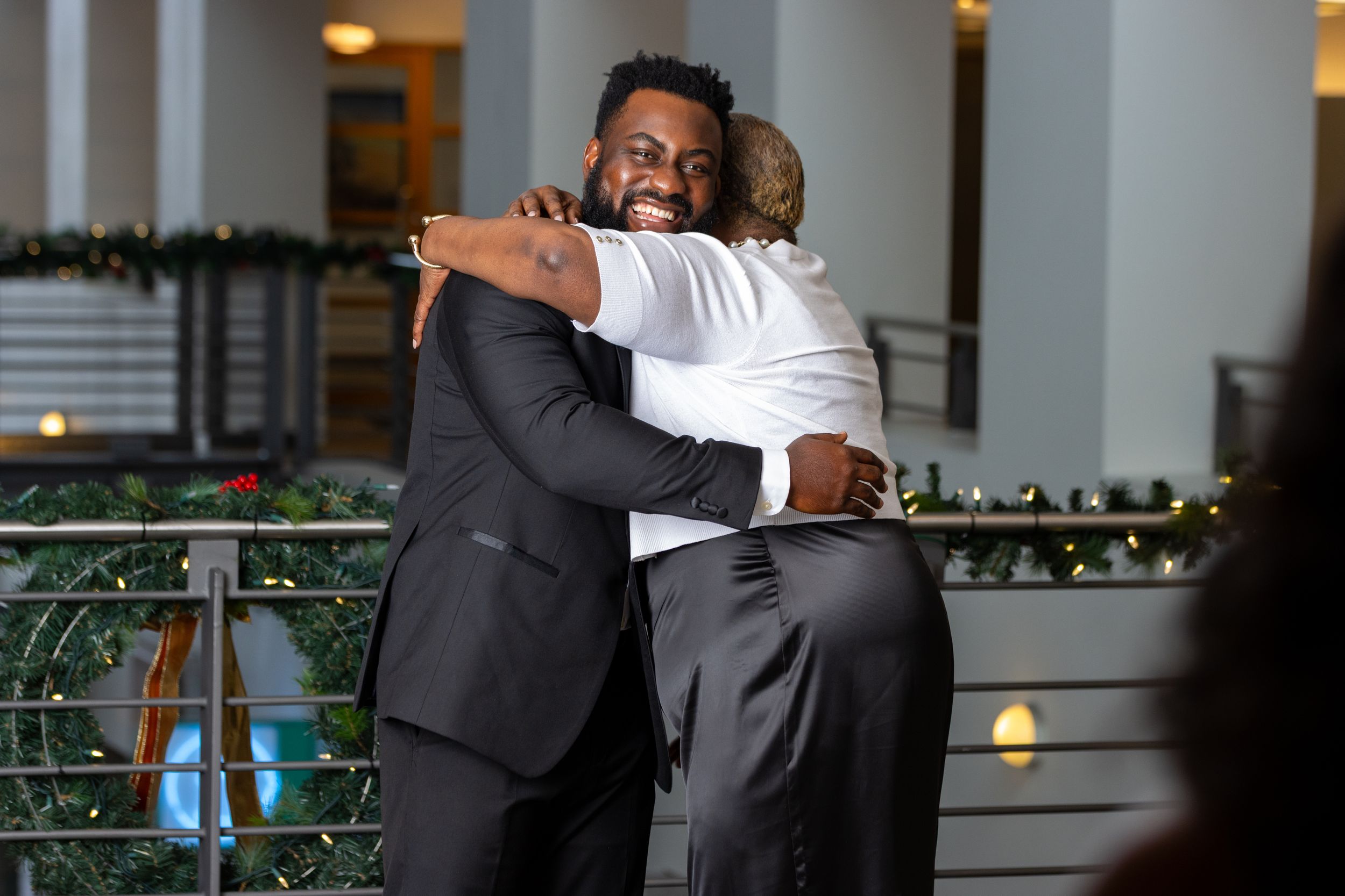 Two people in formal attire share a warm embrace at an indoor venue decorated with twinkling lights and greenery.