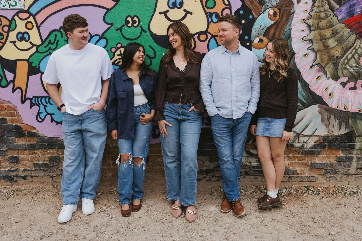 Group of people standing casually against a colorful graffiti wall wearing jeans and casual attire.