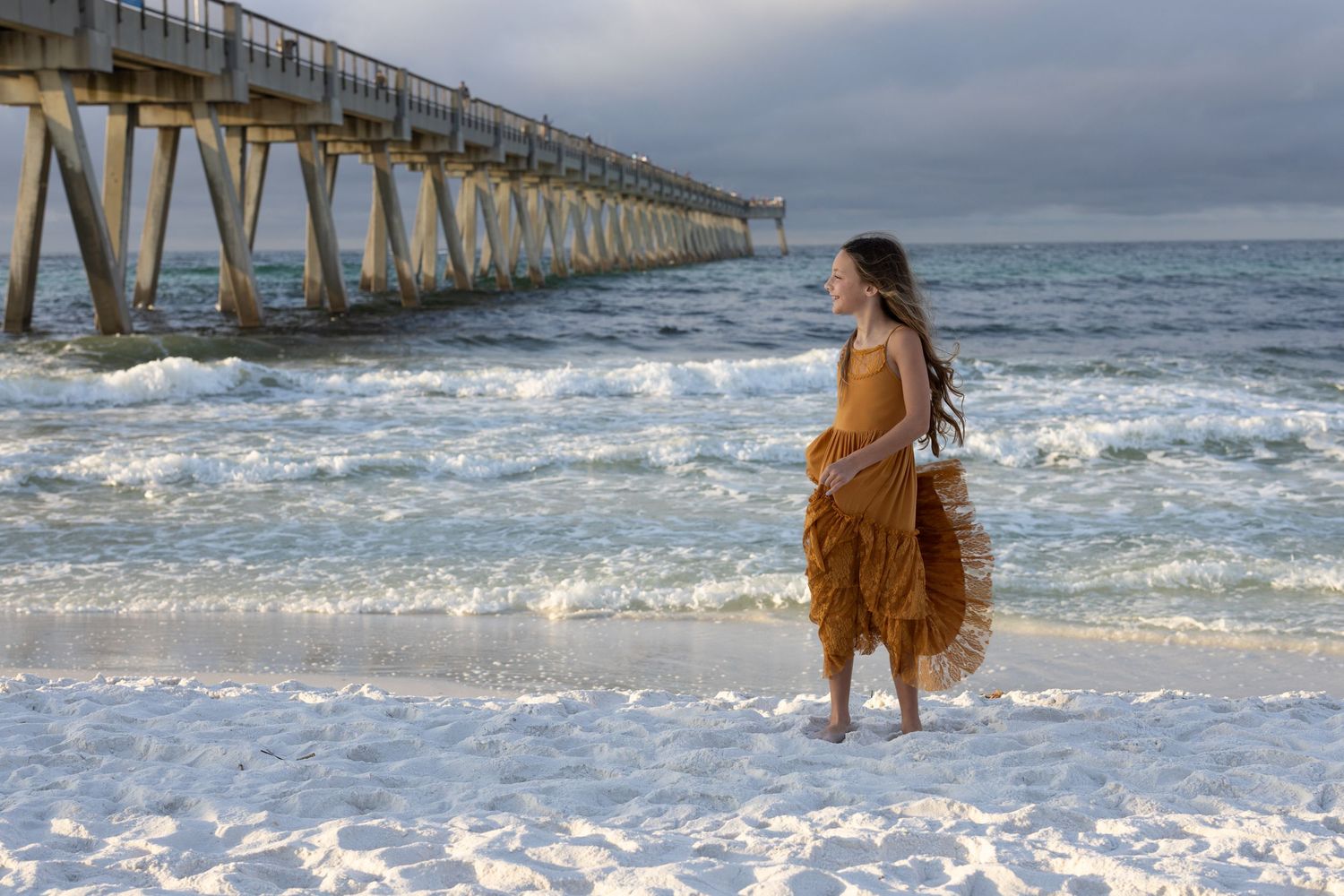 A person in a flowing dress walks along a beach pier during sunset creating a dreamy coastal scene.