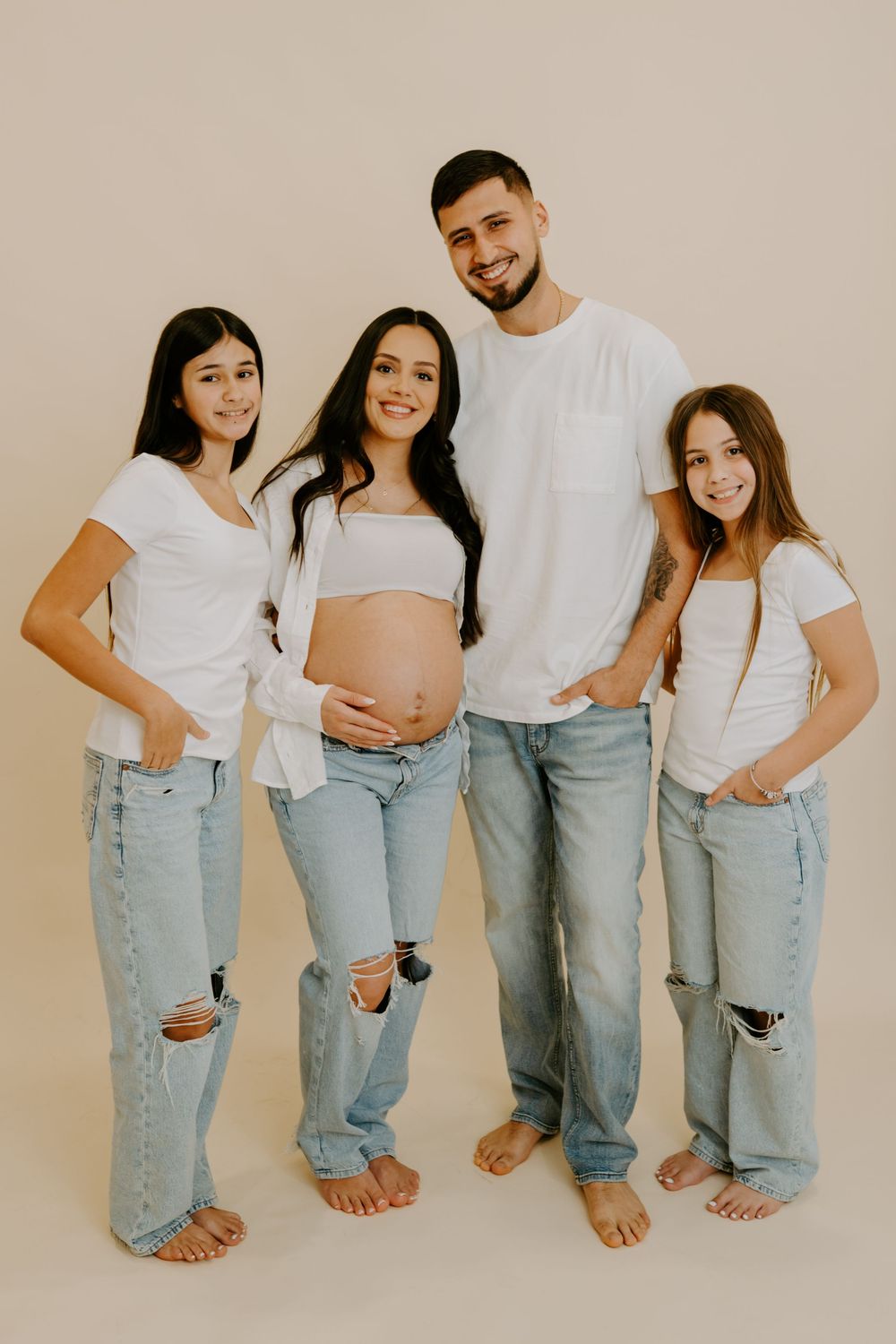 Family wearing matching white shirts and light denim jeans pose together showing their legs and feet against neutral wall.