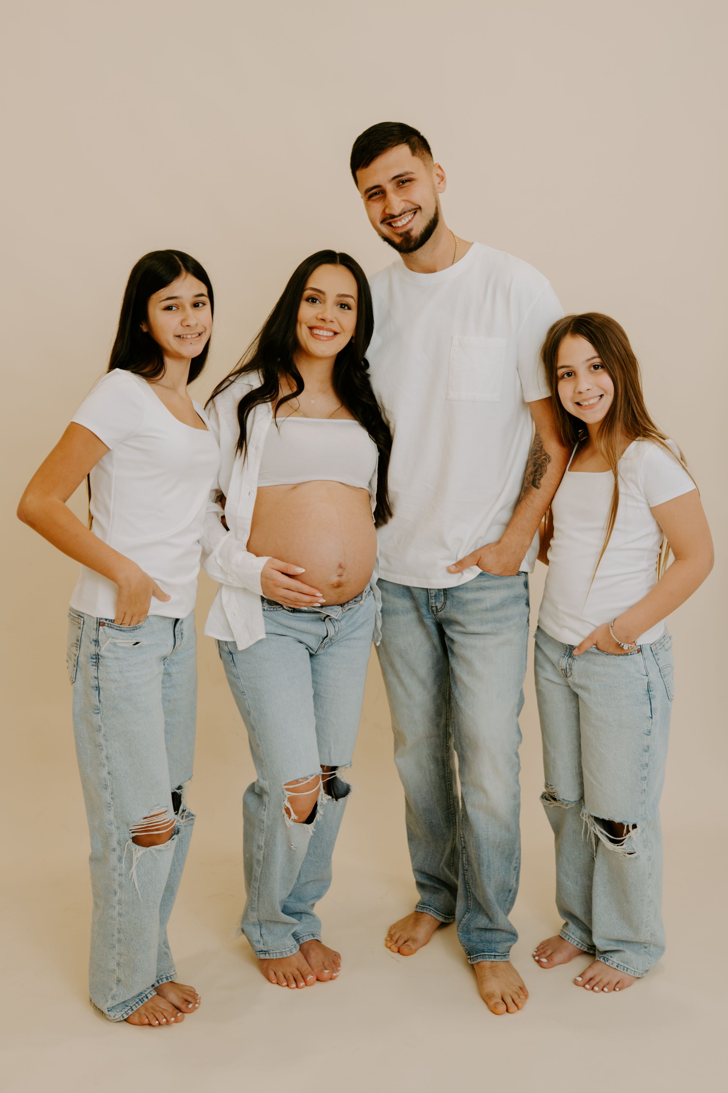 Family wearing matching white shirts and light denim jeans pose together showing their legs and feet against neutral wall.