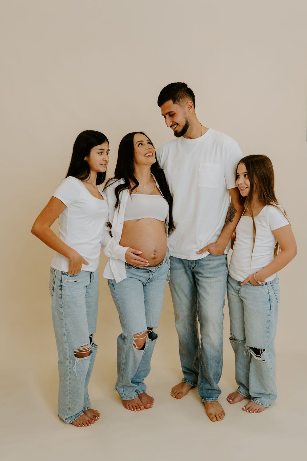 A family group wearing matching white tops and ripped blue jeans poses barefoot against a neutral backdrop.