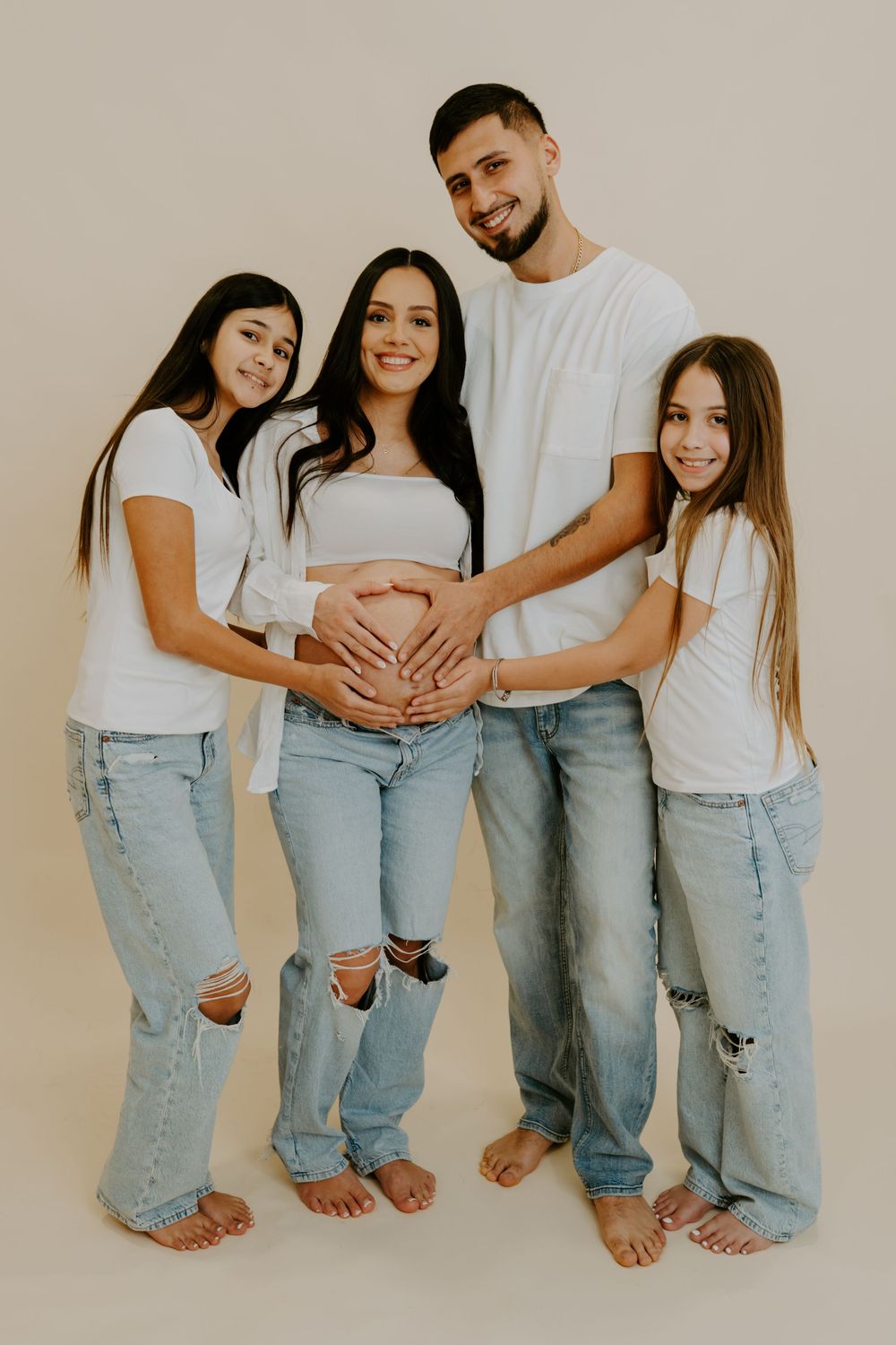 A family wearing white tops and ripped jeans poses barefoot against beige background while touching a pregnant belly.