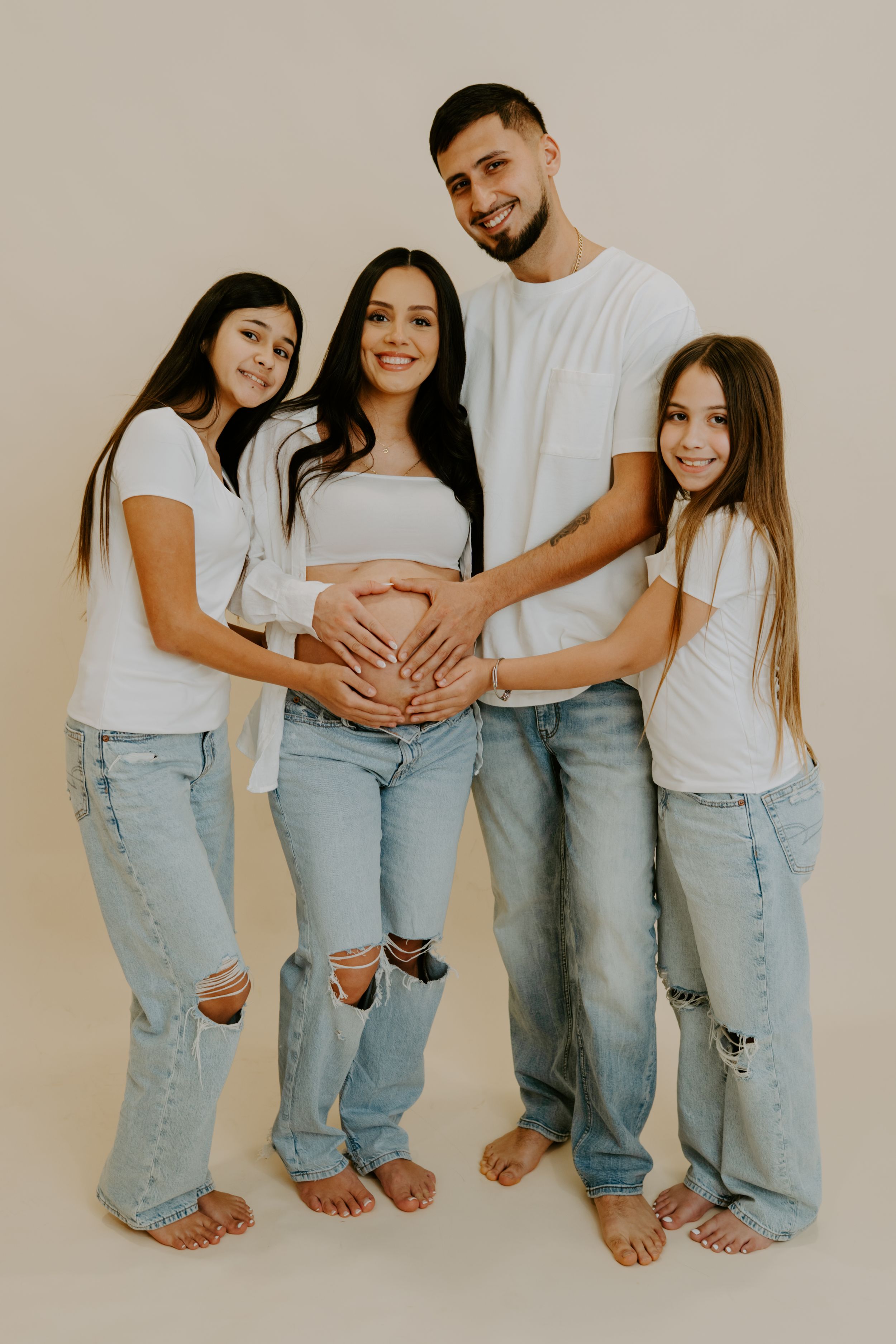 A family wearing white tops and ripped jeans poses barefoot against beige background while touching a pregnant belly.