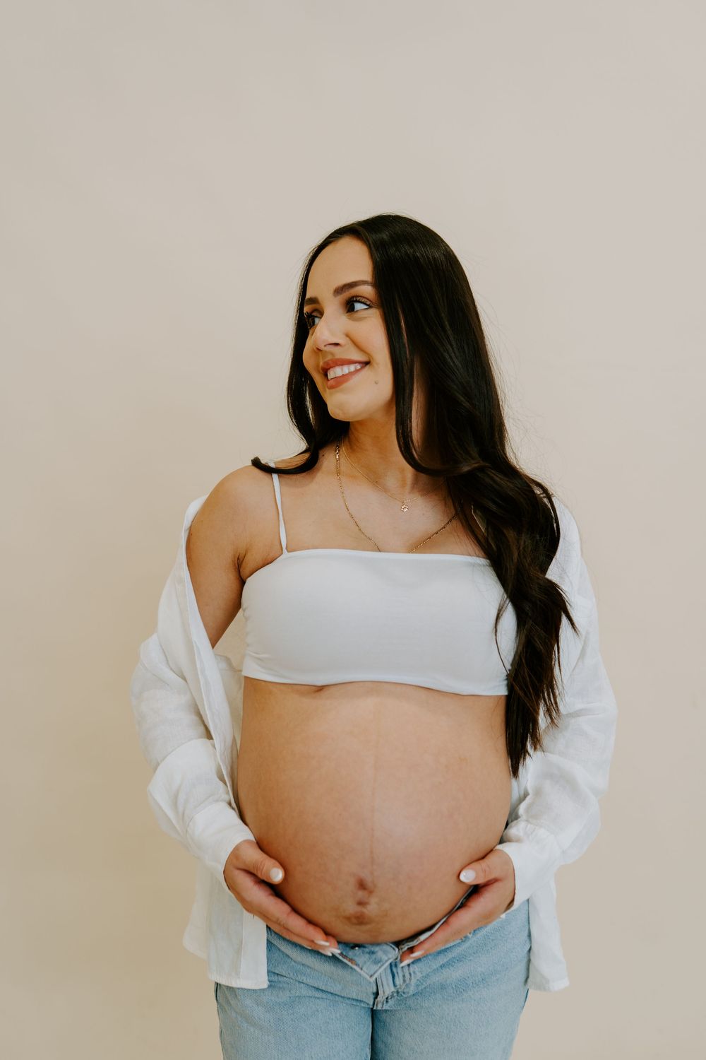 A maternity photoshoot showing a pregnant woman in a white crop top and jeans against a neutral background.