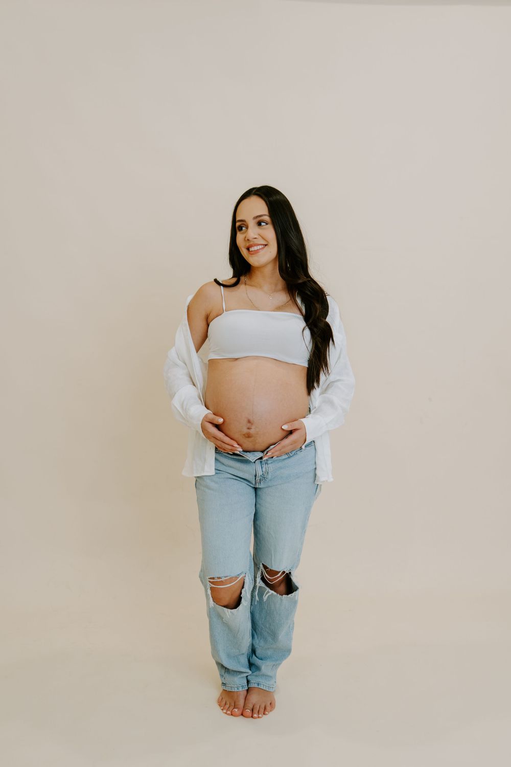 Minimalist maternity photo series showing pregnant woman in light jeans and crop top against neutral backdrop.