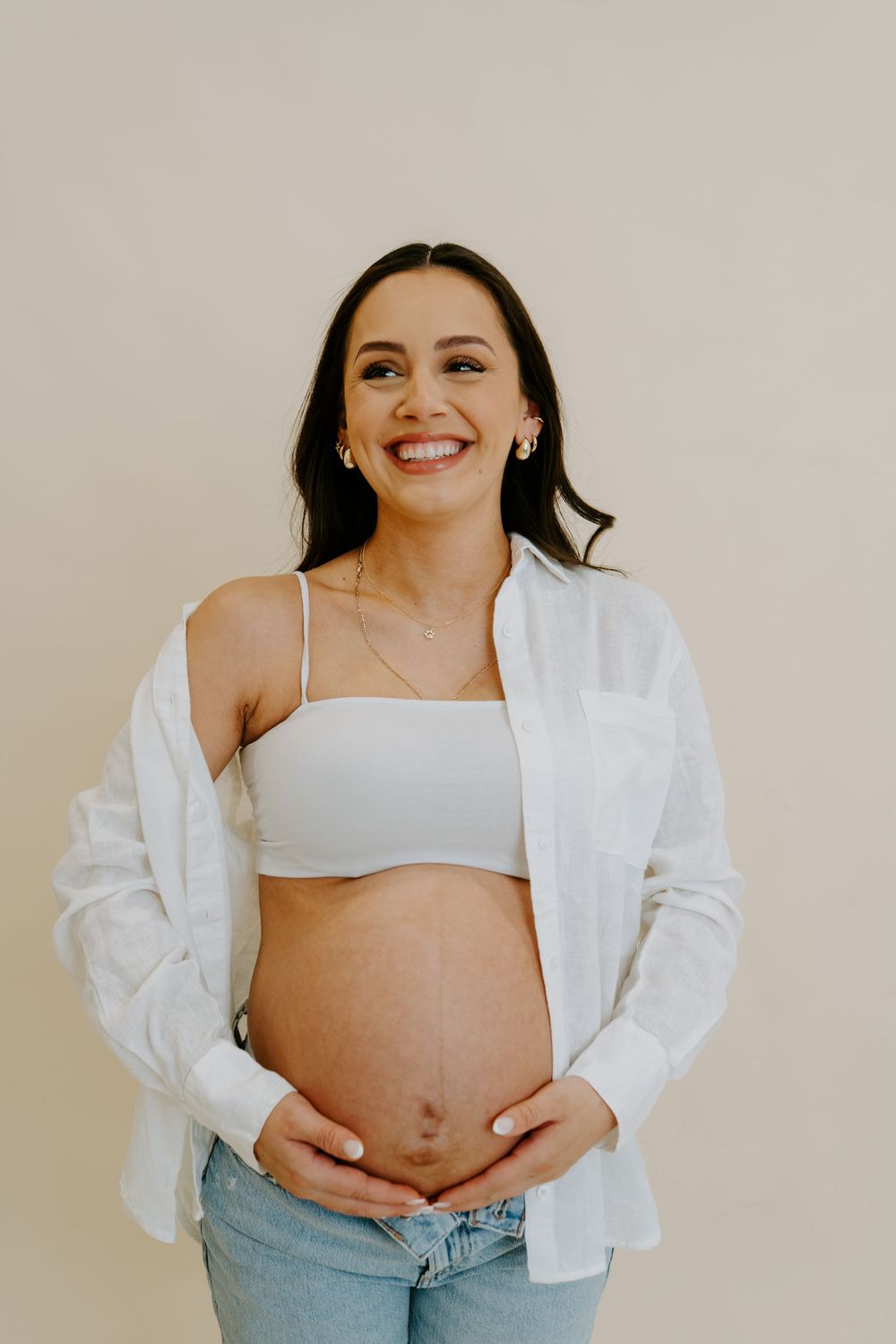 An expectant mother wearing a white shirt and jeans poses playfully in a minimalist studio setting.