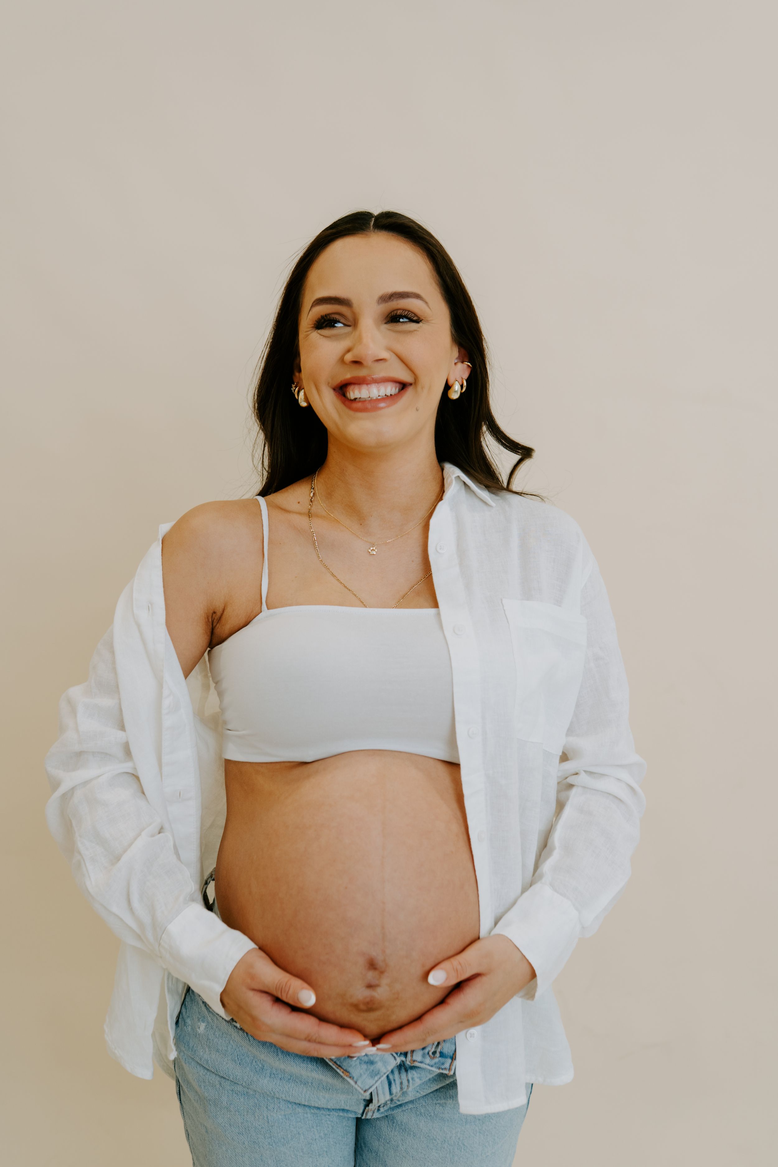 An expectant mother wearing a white shirt and jeans poses playfully in a minimalist studio setting.