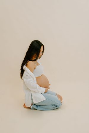 Minimalist maternity photo series showing a woman in white top and jeans sitting against beige backdrop.