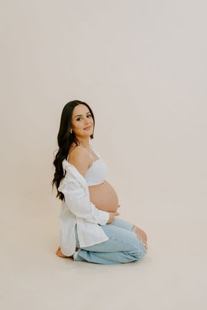 Expectant mother in white top and jeans poses gracefully on floor during maternity photoshoot.