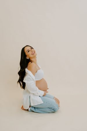 Joyful maternity photo session with woman in casual white top and denim against light backdrop.