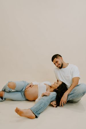 A couple poses together on the floor during an intimate maternity photoshoot in casual attire.
