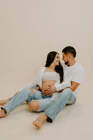 A couple in white t-shirts and jeans cuddle together on a beige background in a minimalist maternity photoshoot.