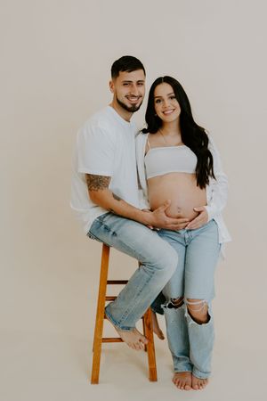 A couple sits intimately on a wooden stool wearing casual attire of white tops and ripped jeans in a minimalist setting.