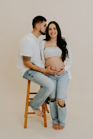A couple shares an intimate moment while sitting on a wooden stool during a photo shoot.