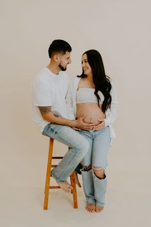 Casual maternity session with couple in denim sitting on wooden stool.