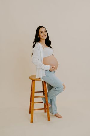 Minimalist maternity photoshoot featuring a person in a white top and jeans sitting on a wooden stool against beige backdrop.