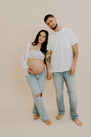 Couple in matching white tops and light denim pose barefoot for casual maternity photos.