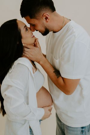 Intimate maternity photoshoot with couple in white clothing sharing tender moments against neutral background.