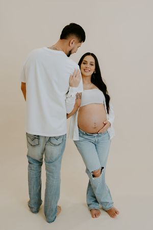 Modern maternity photoshoot featuring couple in white and denim against light backdrop.