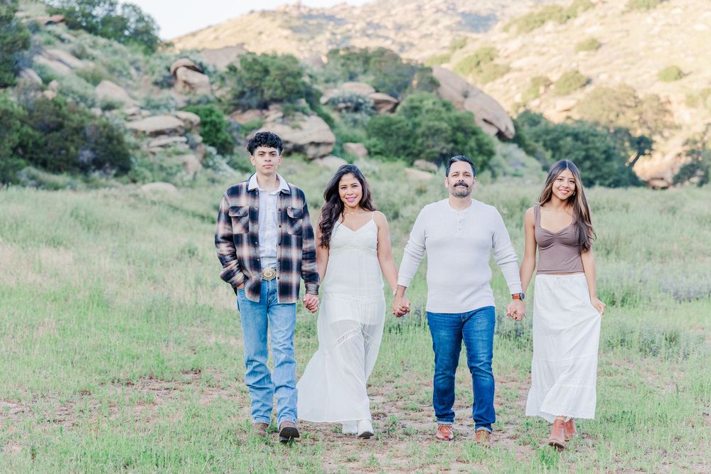 A group of four people stand together holding hands in a scenic grassy hillside setting on a sunny day.