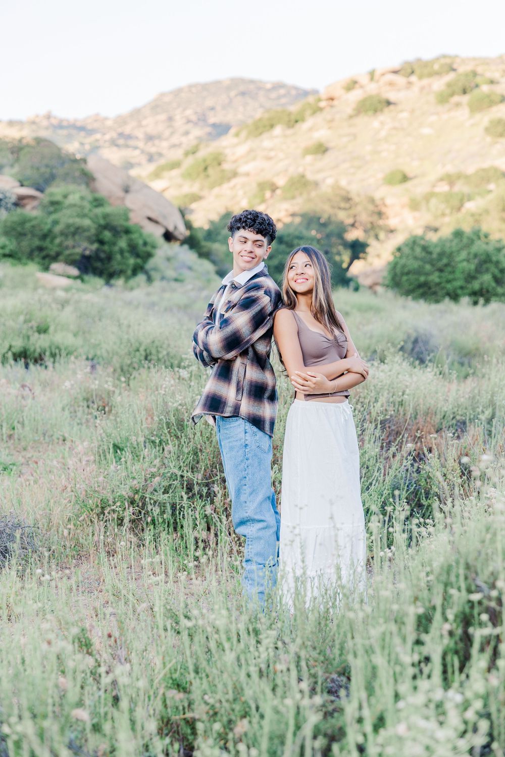 A couple embraces in a field of tall grass and wildflowers with rolling hills in the background during golden hour.