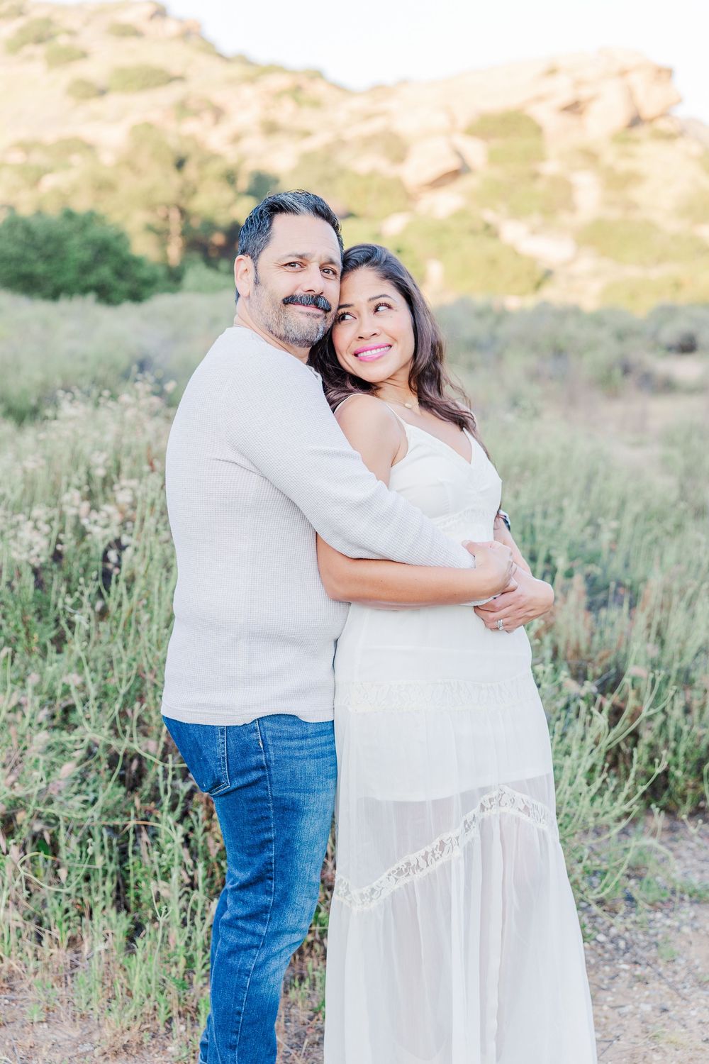 A couple embraces outdoors in a natural setting with mountains and wildflowers in the background.