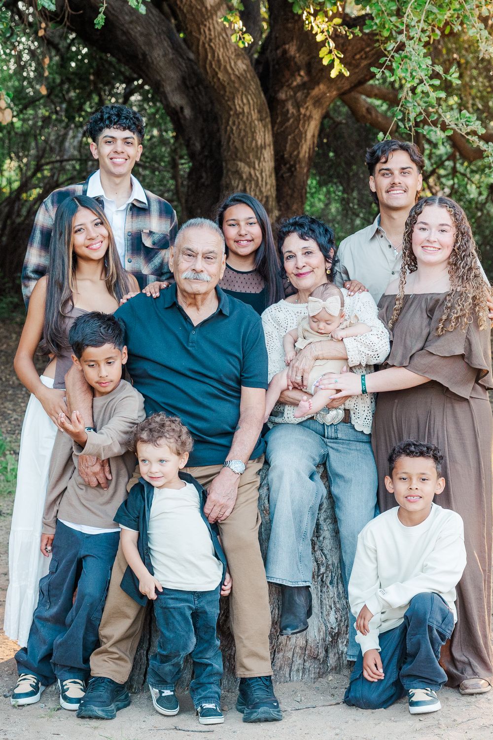Extended family group poses together under a large tree in coordinated neutral-toned casual outfits.