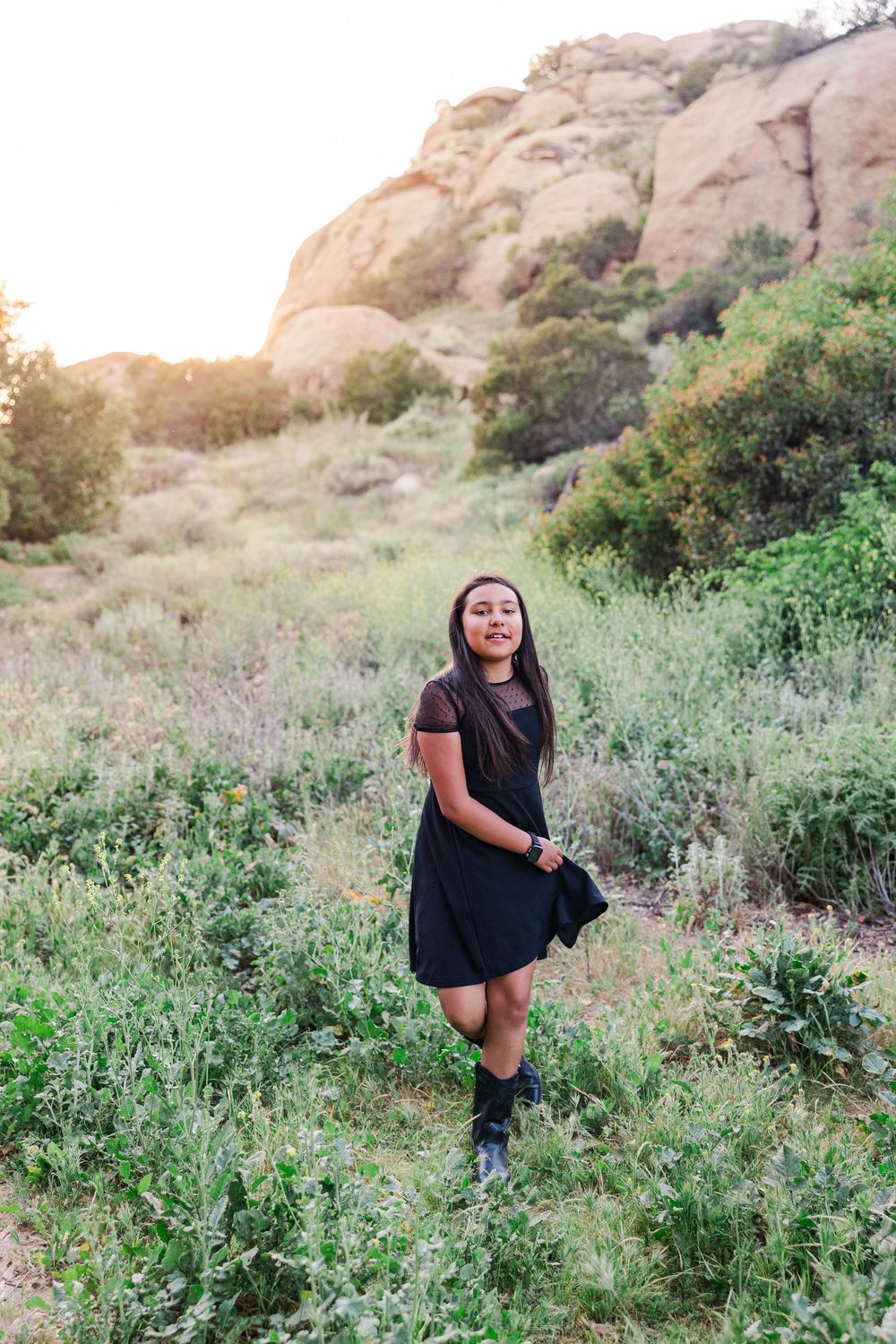 A person in a black dress stands on a hillside trail with large rock formations and green vegetation at sunset.
