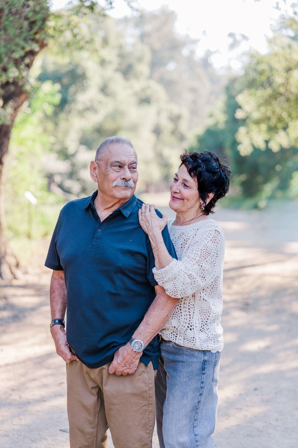 Elderly couple shares tender moments while walking together on a dirt path in a sun-dappled outdoor setting.