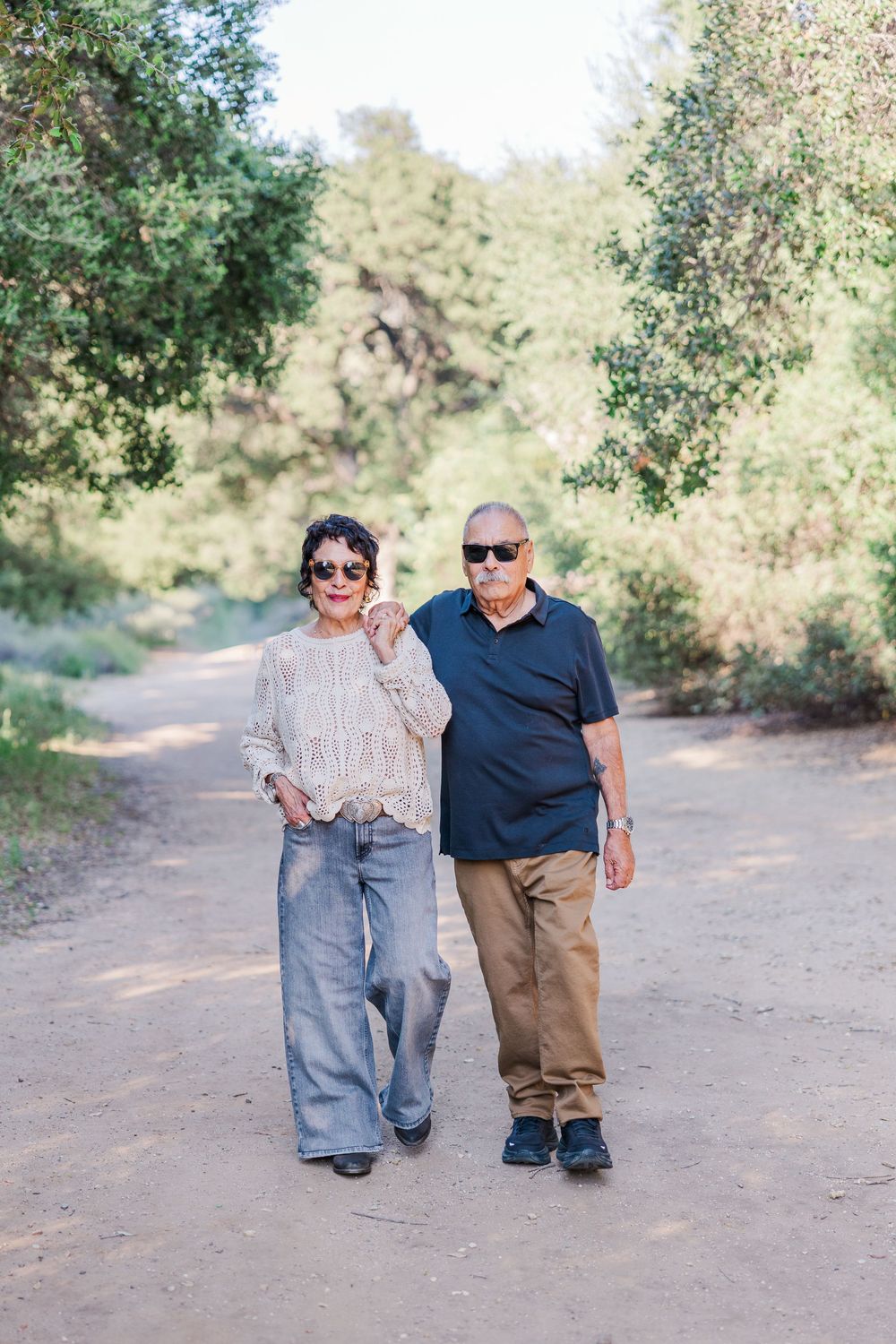 Two people walking together on a dirt path surrounded by green trees on a sunny day.