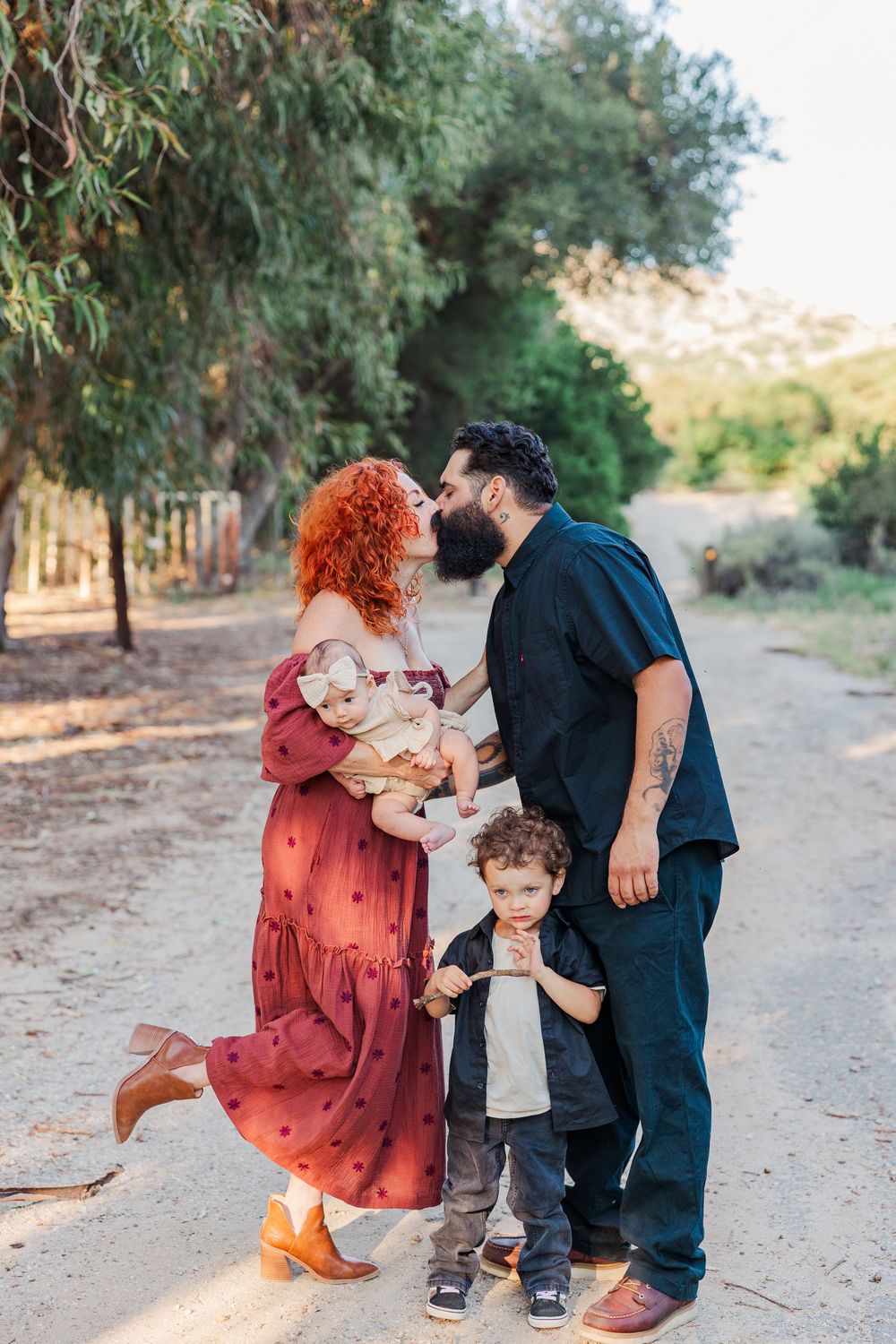Family in coordinating outfits shares tender moments together on a dirt path surrounded by trees.