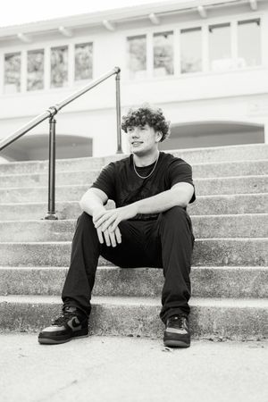Black and white portrait of someone sitting casually on concrete steps wearing dark clothing.