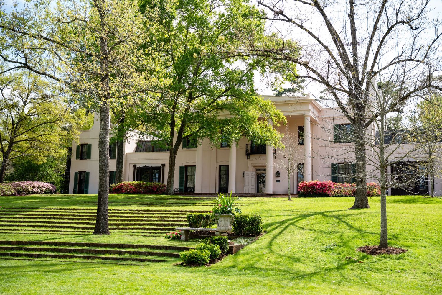 A grand white neoclassical estate home surrounded by lush green lawns, mature trees, and blooming azalea shrubs in spring.