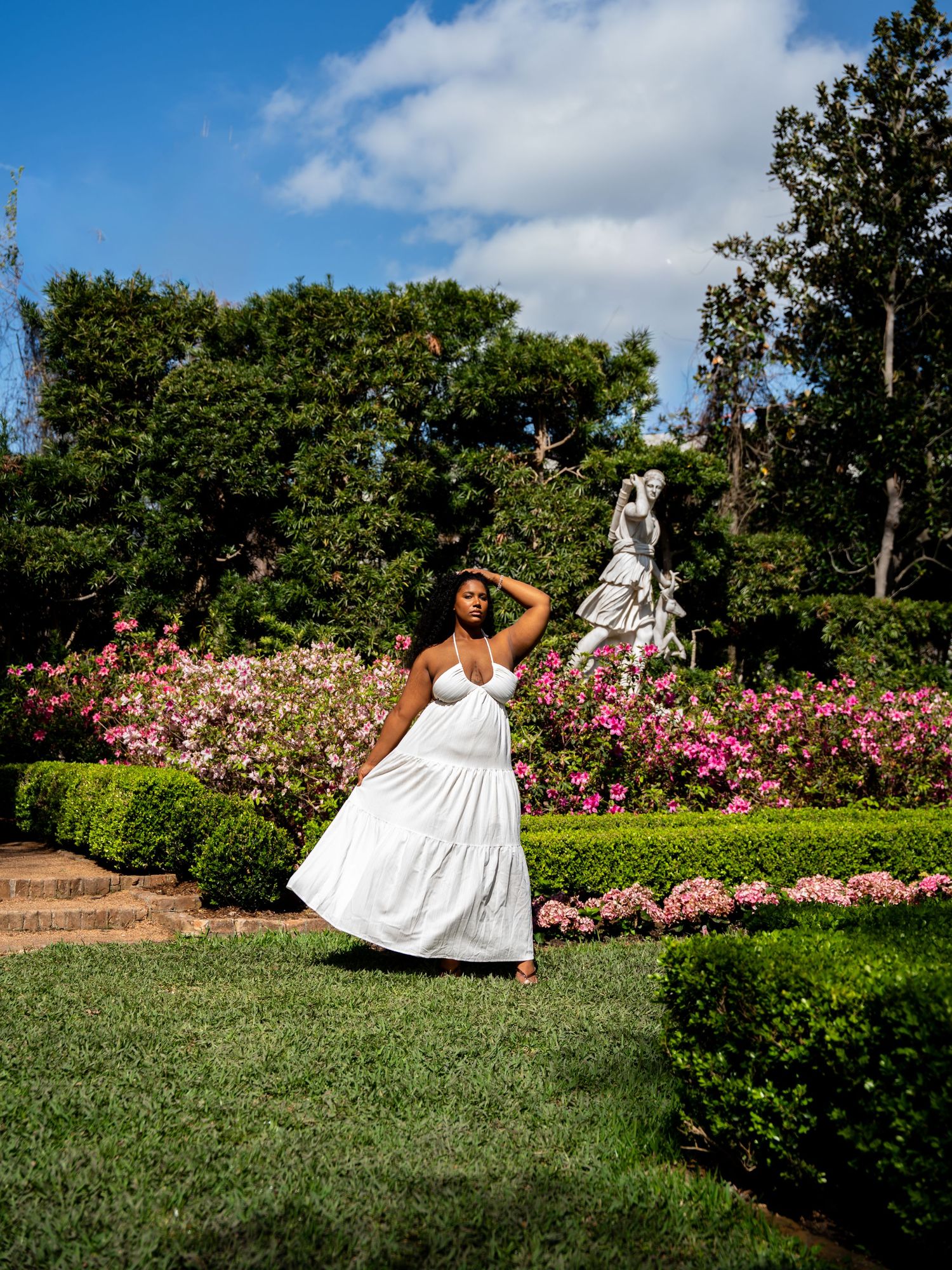 Woman in white maxi dress posing in a lush garden with pink flowers and a classical statue.
