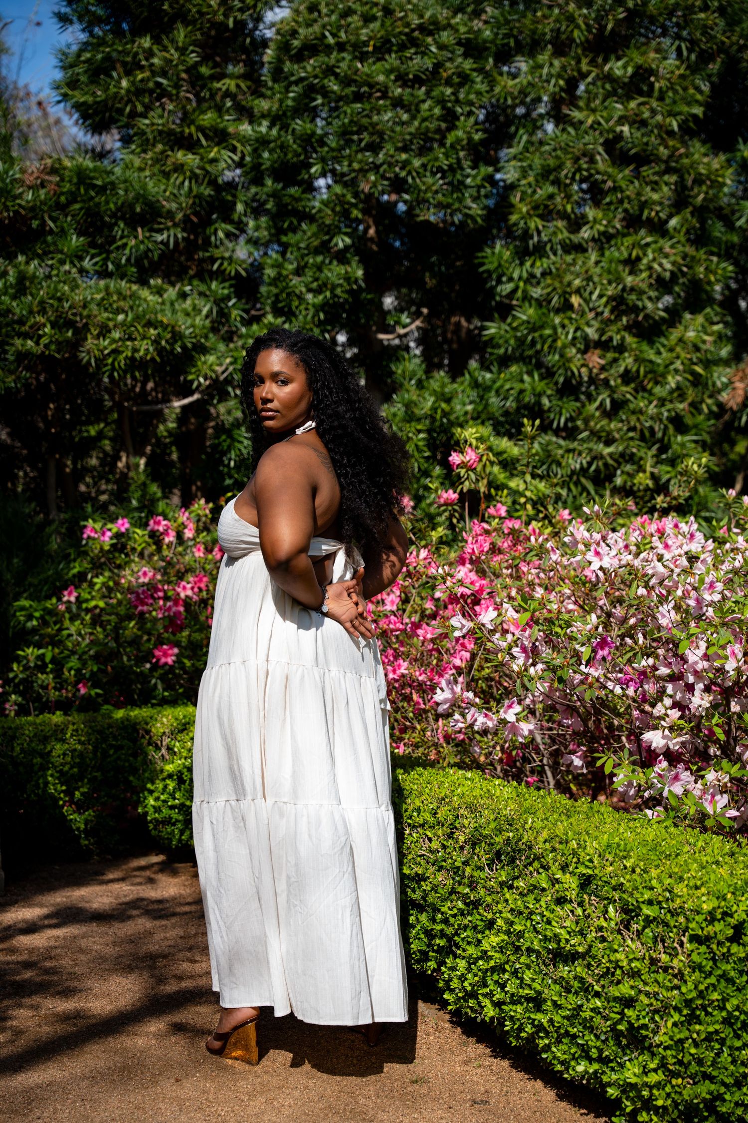 Black woman in white tiered maxi dress standing before vibrant pink azalea bushes in a garden.