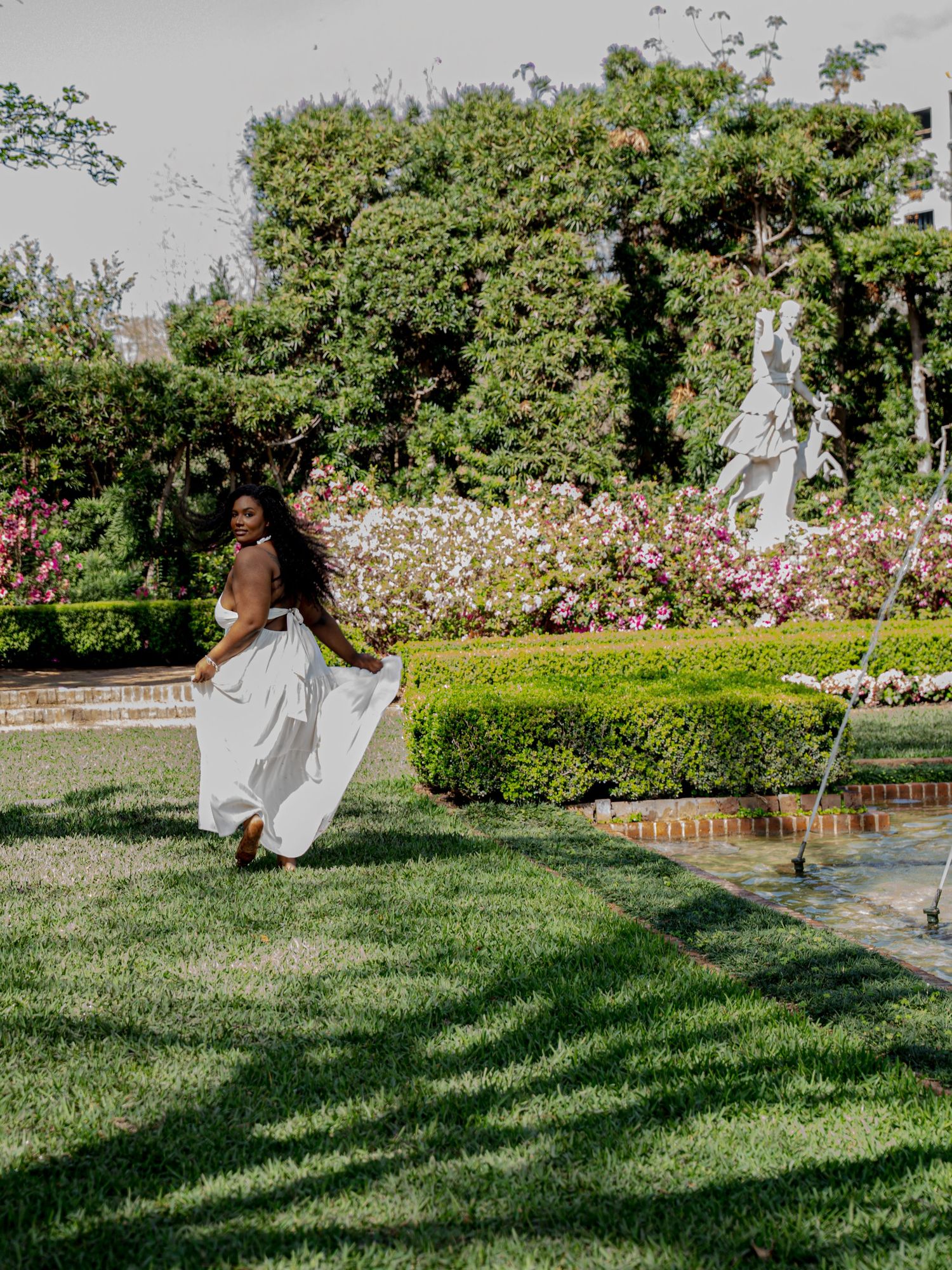 Woman in flowing white dress running across green lawn in a formal garden with statues and flowers.