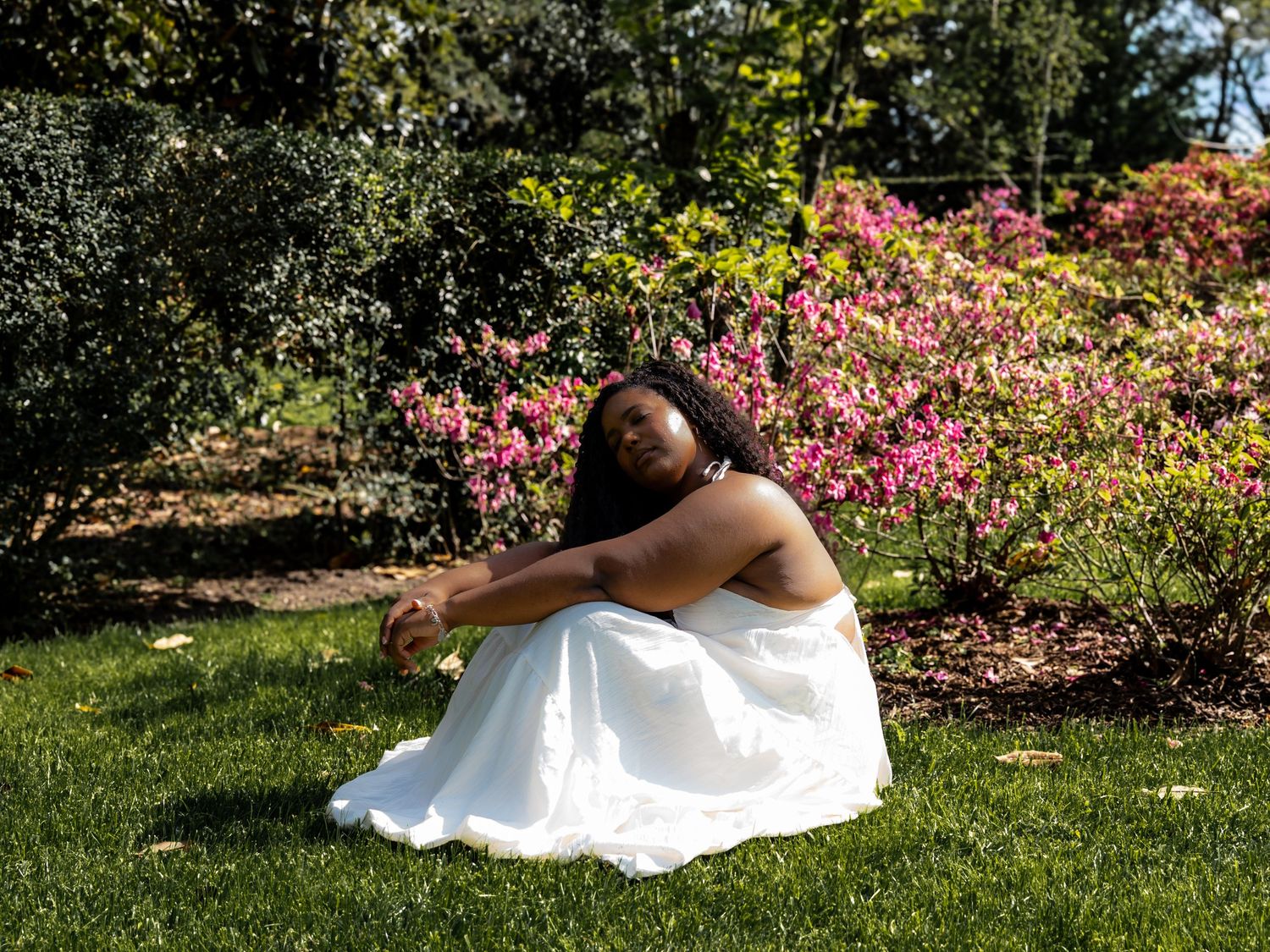 Woman in white dress sitting on grass surrounded by colorful blooming flowers in a sunny garden.