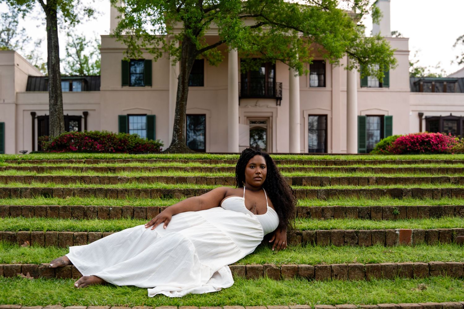 Woman in white gown reclining on moss-covered steps before a grand columned estate building.